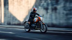 a man riding on the back of an orange motorcycle down a street next to a stone wall