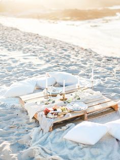 a table set up on the beach with candles and plates in front of it, ready to be served