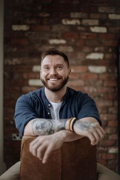 a man sitting on top of a brown chair with his arms crossed and tattoos on his arm