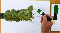 a person holding a paintbrush in front of a painting with water and trees on it