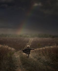 a person walking down a dirt road in the middle of a field with a rainbow behind them