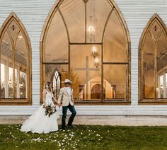 a bride and groom standing in front of a church with large arched windows, dressed in white