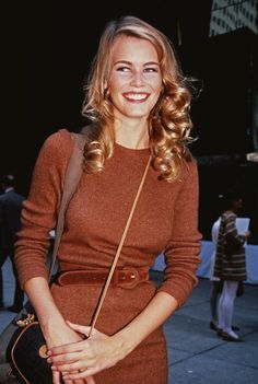 a woman in a brown dress is smiling and holding a handbag on the street