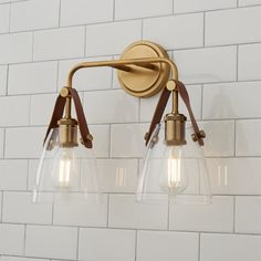 two lights are attached to the wall in front of a white brick wall, one is gold and the other is clear