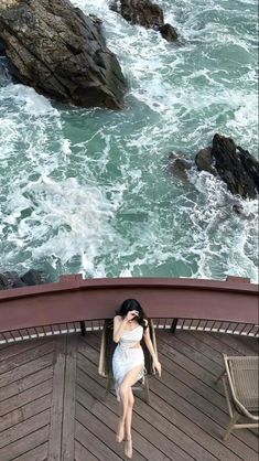 a woman in a white dress standing on a wooden deck next to the ocean and rocks