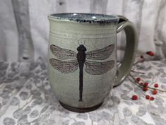 a mug with a dragonfly on it sitting on a table next to some red berries