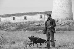 a man standing next to a wheelbarrow with a lighthouse in the back ground