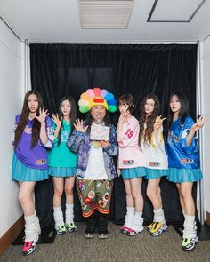 an old man standing in front of a group of young women wearing colorful outfits and holding up their hands