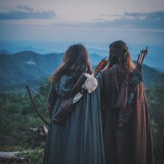 two women dressed in medieval clothing look out over the hills and mountains with bows on their heads