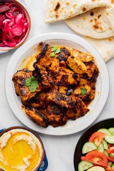 a white plate topped with chicken next to bowls of vegetables and pita bread