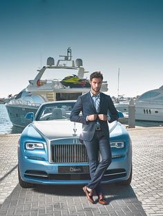 a man in a suit standing next to a blue rolls royce parked near a boat