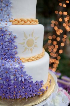 a three tiered cake with purple flowers on the bottom and gold trimmings