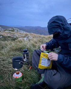 a man sitting on top of a grass covered hill next to a camping stove and cup