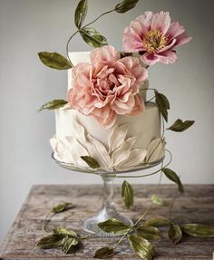 a white cake with pink flowers on top and green leaves around the edges, sitting on a wooden table