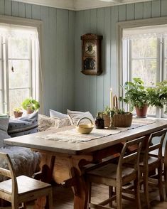 a dining room table and chairs in front of two windows with potted plants on them