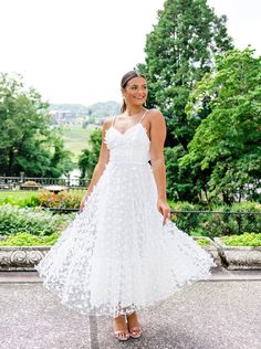 a woman wearing a white dress standing in front of some trees and bushes with her hands on her hips