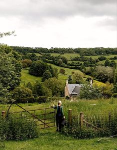 an old woman standing in front of a gate on the side of a lush green hillside