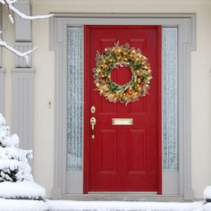 a red door with a wreath on it in front of snow covered bushes and trees