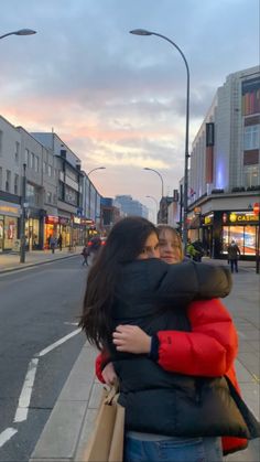 two women hugging each other on the street in front of shops and buildings at dusk