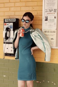 a woman standing next to a pay phone holding a cup and wearing sunglasses with her hands on her hips