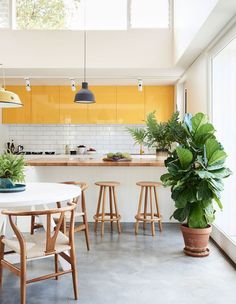 a kitchen with yellow cabinets and wooden stools next to a white dining table surrounded by potted plants