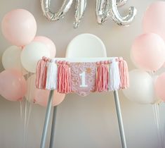 a baby's first birthday party with pink and white balloons, tasselled chair