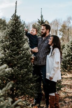 a man and woman holding a baby in front of a christmas tree while standing next to each other