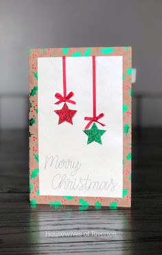 a christmas card with two red and green stars hanging from it's sides on a wooden table