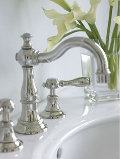 a white sink with two faucets and some yellow flowers in the vase next to it