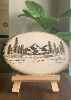 a wood slice with trees and mountains on it is sitting on a table next to a potted plant