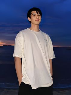 a young man standing in front of the ocean at night wearing a white shirt and black pants