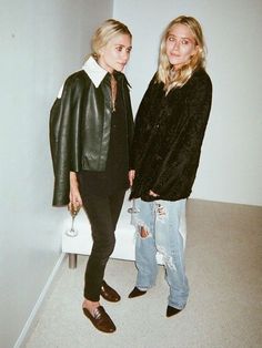 two women standing next to each other in front of a white wall and one is wearing a black leather jacket