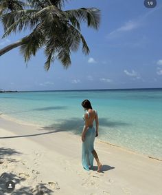 a woman in a long dress walking on the beach next to a palm tree and clear blue water