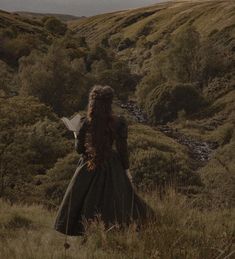 a woman with long hair is standing in the grass and looking down at her book