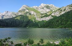 a lake surrounded by mountains and trees in the foreground with green foliage on either side