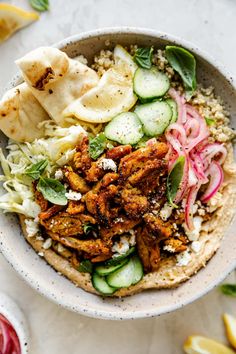 a bowl filled with meat and vegetables next to sliced lemons, cucumbers, and pita bread