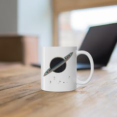 a white coffee mug sitting on top of a wooden table next to a laptop computer