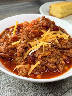 a white bowl filled with chili and cheese next to a piece of bread on top of a wooden table