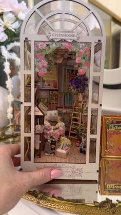 a person holding up a miniature dollhouse in front of a table with flowers on it