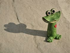 a green toy frog with eyes wide open sitting on the ground next to a shadow