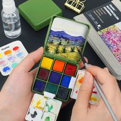 a person is holding a paintbrush and some watercolors on a table with other items