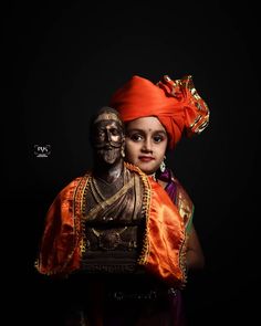 a woman in an orange turban and headdress poses with a statue