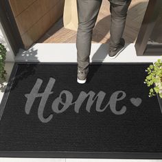 a person standing on the front door mat that says home in grey letters, with plants and potted plant beside it