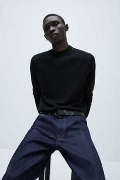 a man sitting on top of a chair wearing jeans and a black sweater with his hands in his pockets