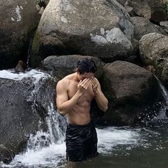 a shirtless man standing in the water next to some rocks and holding his hands to his face