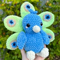 a hand holding a blue and green crocheted stuffed animal in front of some bushes