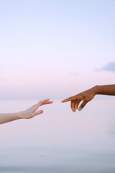 two hands reaching out towards each other in front of a body of water at sunset