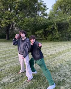 two young men standing on top of a grass covered field with trees in the background