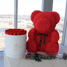 a red teddy bear sitting next to a white box with roses in it and a large window behind it