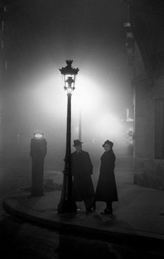 two people standing next to a street light on a foggy night in black and white